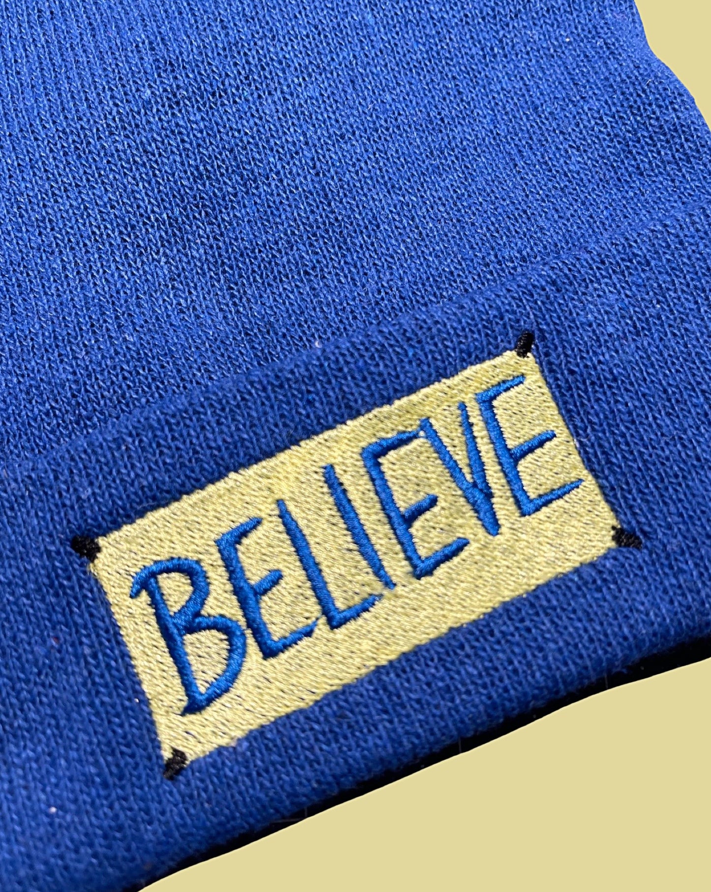 detail view of the embroidered "believe" sign from Ted Lasso on royal blue knit beanie