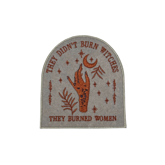 Embroidered "They Didn’t Burn Witches" patch with mystical hand design in rust and gold on a gray background.
