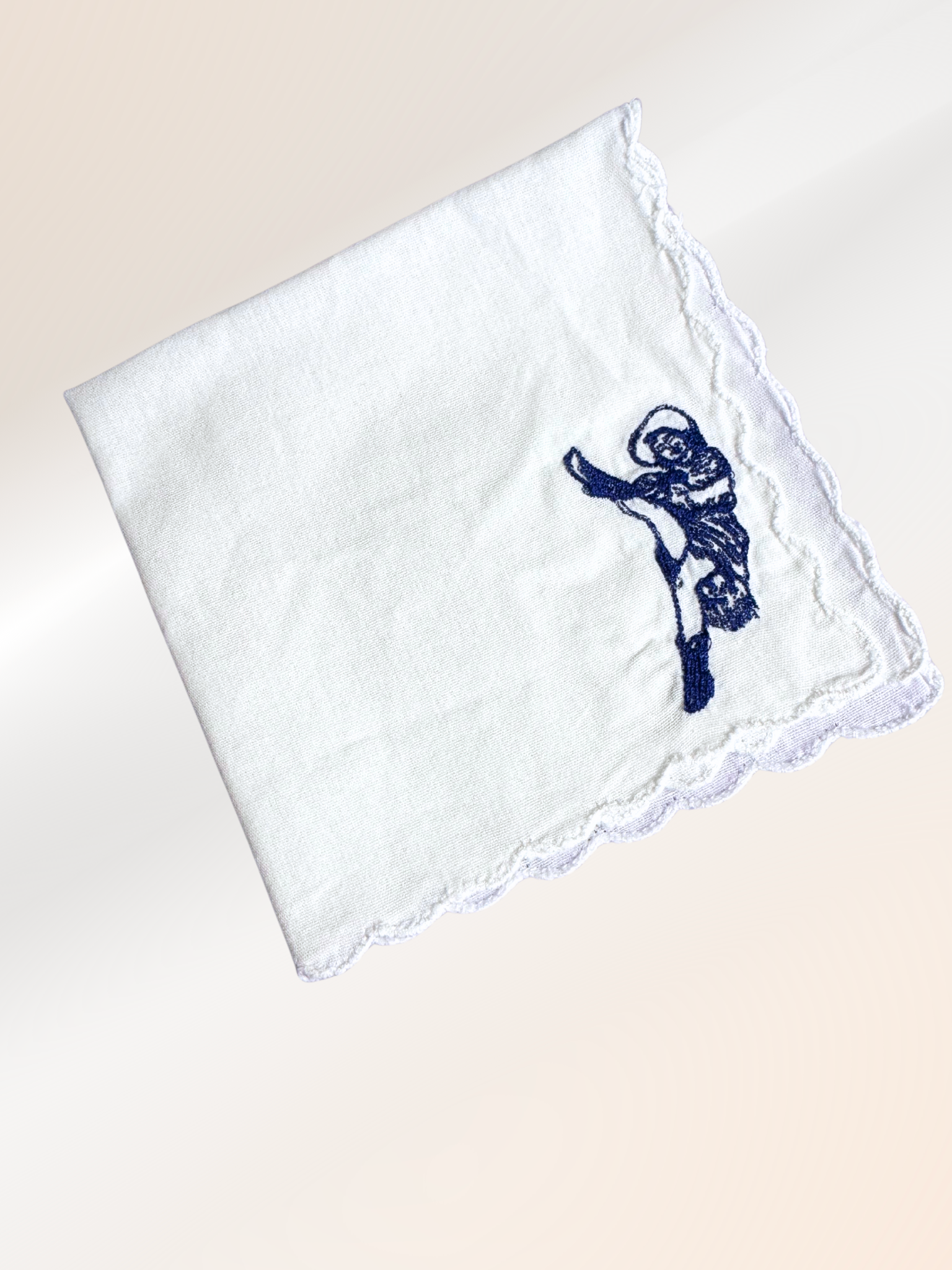A folded embroidered handkerchief showcasing a vintage French cabaret dancer stitched in blue on soft white fabric, handmade in Kansas City.


