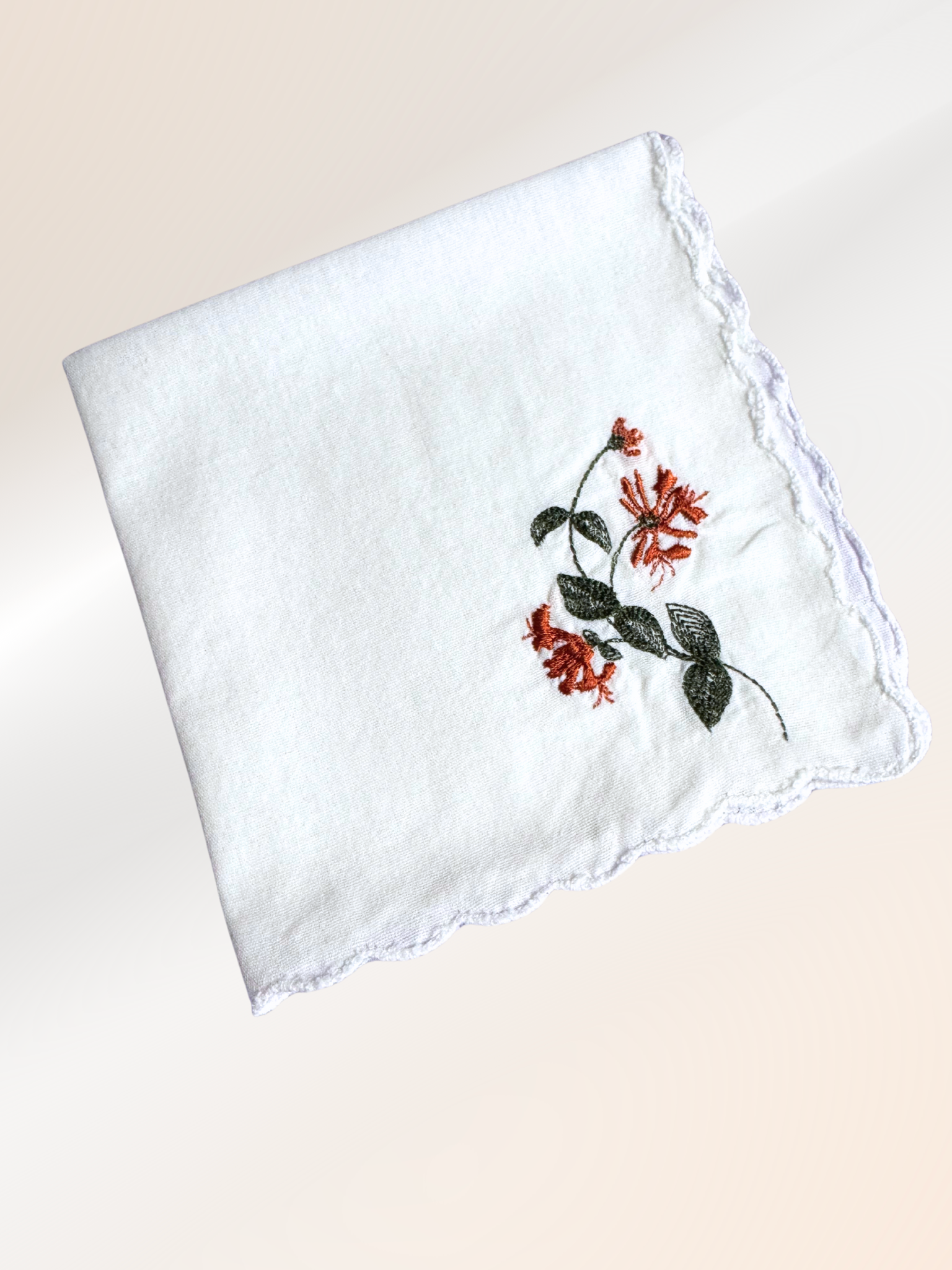 A folded embroidered handkerchief showcasing a delicate honeysuckle vine stitched in rust and green on soft white fabric, handmade in Kansas City.

