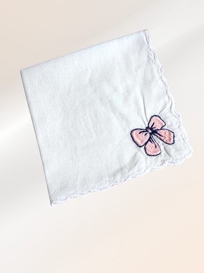 A folded embroidered handkerchief showcasing a delicate ribbon bow stitched in blush pink and navy on soft white fabric, handmade in Kansas City.

