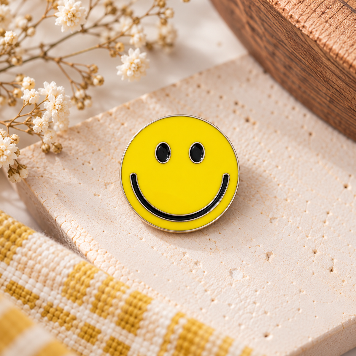 Bright yellow smiley face enamel pin with black eyes and smile and silver-tone outline, styled on cream stone