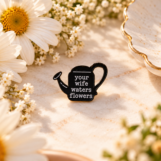 Black watering can enamel pin reading your wife waters flowers, styled with white daisies; inspired by Taylor Swift's thanK you aIMee.