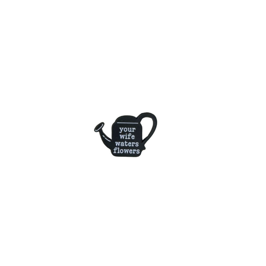 A black enamel pin shaped like a watering can with the words “your wife waters flowers,” referencing a Taylor Swift lyric.